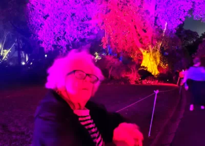 An elderly woman sits in a wheelchair in front of a tree lit up in a blaze of purple, red and yellow light