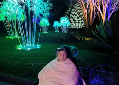 A smiling woman sits in a wheelchair wrapped in a blanket in front of trees lit up in bright green.