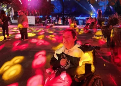 A man sits in a wheelchair and smiles, surrounded and bathed in multi-coloured lights.