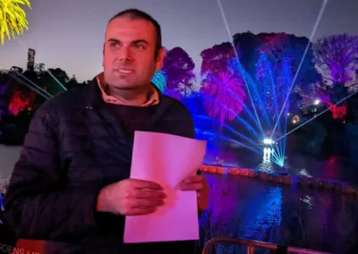 A man stands smiling and holding a piece of paper, with water fountains and trees lit up blue and purple behind him.