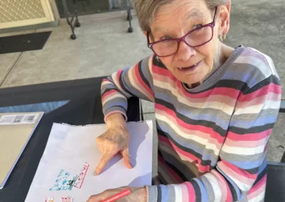 A seated elderly woman shows the drawing she’s working on