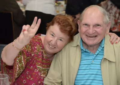 A smiling older woman gives a peace sign with her hand beside a smiling older man,