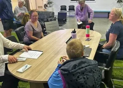 Three people sit around a table while a fourth, elderly woman, stands.