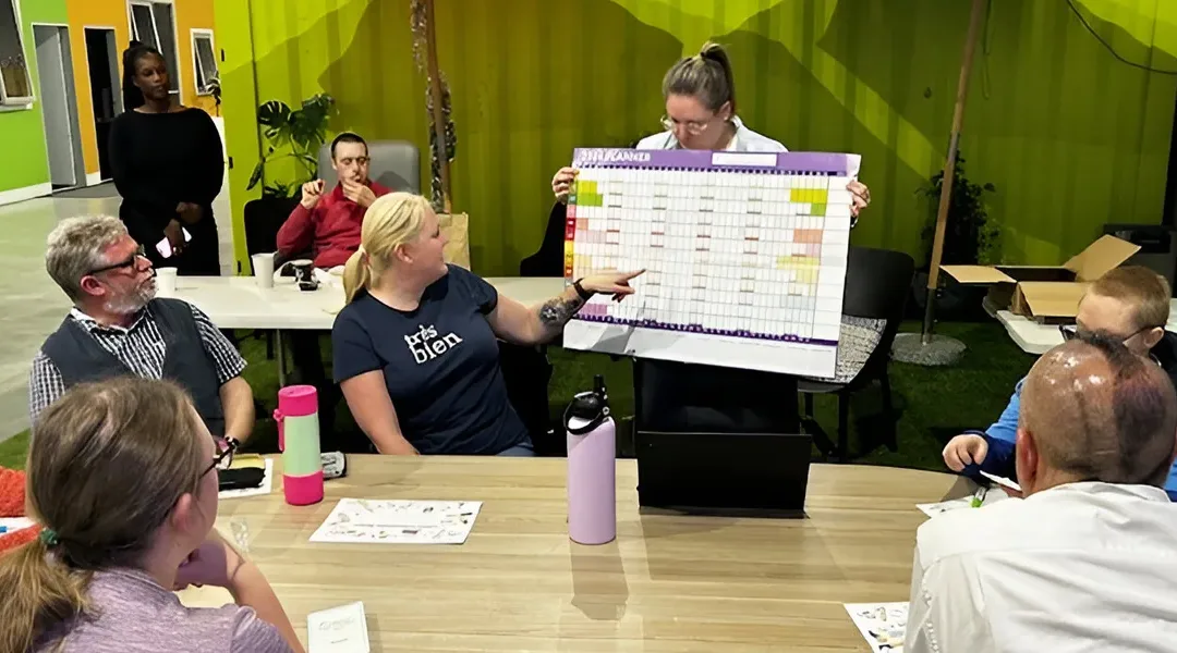 A woman stands with a large calendar board while people seated at a table – and behind – look on